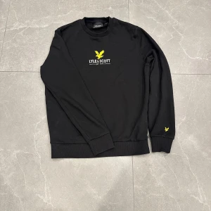 Svart sweatshirt från Lyle & Scott - Svart sweatshirt från Lyle & Scott med klassisk gul logga på bröstet och ärmen. Tröjan har rund hals, ribbade muddar och är tillverkad i mjuk bomull. Perfekt för en clean och stilren look.