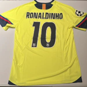 Barcelona Ronaldinho 10 bortatröja Nike - Gul Barcelona bortatröja med Ronaldinho och nummer 10 på ryggen. Tröjan har korta ärmar, mörkblå detaljer och klubbmärke på bröstet. Champions League-märke på ärmen och klassiska Nike-loggan. Tillverkad i lätt och ventilerande material. Replika. Endast handtvätt gäller!