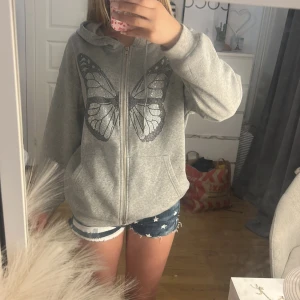 Grå kofta med fjärilstryck - Grå hoodie med huva och dragkedja framtill. På bröstet finns ett stort fjärilsmotiv i svarta och silvriga stenar som ger en snygg effekt. Tröjan har känguruficka och långa ärmar, perfekt för en chill och trendig look. Tyvärr så är det svårt att se vilken storlek det är, men den passar mig som är 165cm. 
