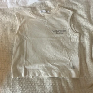 Beige ärmlös topp från Calvin Klein - Snygg beige ärmlös linnetopp från Calvin Klein Jeans med rund halsringning. Toppen har diskret CK-logga nertill och broderad Calvin Klein Jeans-text på bröstet. Perfekt för en clean och stilren look. En liten fläck nera vil CK-loggan, och en liten fläck uppe vid CK-loggan.