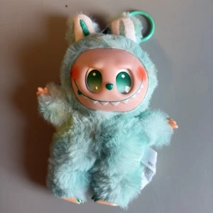 Popmart fluffig mintgrön handväska - Supergullig liten handväska från Popmart i form av ett mintgrönt monster med mjuk päls och stora öron. Väskan har ett plastansikte med gröna ögon och ett brett leende, samt en praktisk ring för att fästa på väskan eller nycklarna.