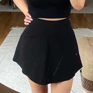 Svart kortkjol från Oiso Design - Enkel och stilren svart kortkjol från Oiso Design. Kjolen har en lätt utställd passform och stängs med dragkedja i sidan. Perfekt att matcha med crop top eller stickad tröja. Tidlös och lätt att styla till olika looks.