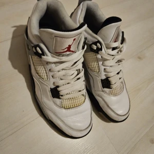 Nike Air Jordan 4 Retro Vit/Röd - Säljer ett par klassiska Nike Air Jordan 4 Retro i vitt skinn med svarta och röda detaljer. Skorna har meshpaneler, snörning och den ikoniska Jumpman-loggan på plösen. Sulan är svart, vit och röd med Nike-logga undertill. Perfekta för dig som gillar streetstyle och sneakerskultur.