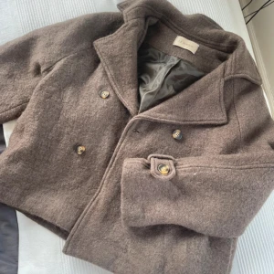 Brun ull kappa från  - Säljer en brun fluffig kappa i ull. Jackan har dubbelknäppning, bred krage och snygga knappar vid ärmsluten. Oversized passform och fodrad insida. 