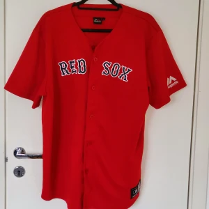 Röd Red Sox baseballskjorta Majestic - Säljer en röd baseballskjorta från Majestic med Red Sox-tryck på bröstet. Skjortan har korta ärmar, knappar framtill och är gjord i polyester. MLB-logga och Majestic-märke finns på ärmen och nertill. Perfekt för dig som älskar sport och vill sticka ut.  Passar M och L
