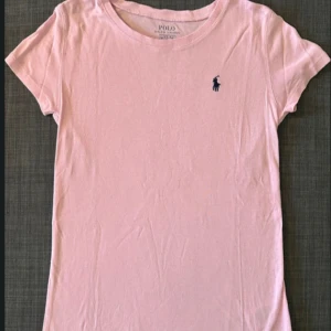 Ljusrosa t-shirt från Polo Ralph Lauren - Snygg ljusrosa t-shirt från Polo Ralph Lauren med klassisk passform och korta ärmar. T-shirten har en liten mörkblå broderad logga på bröstet och är gjord i mjuk bomull. Perfekt basic till garderoben!