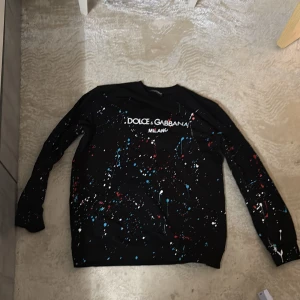 Dolce & Gabbana tröja - Svart långärmad tröja från Dolce & Gabbana med coola färgstänk i vitt, blått och rött över hela plagget. Stor logga framtill. Perfekt statement piece för dig som vill sticka ut med en unik och trendig look.