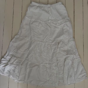 Vit långkjol med volanger - Säljer en flowy vit långkjol med flera volanglager och resår i midjan. Kjolen är luftig och perfekt för varma dagar, enkel att styla med crop tops eller stickade tröjor. Passar dig som gillar en romantisk och avslappnad look.