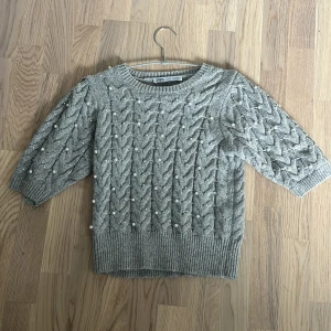 Grå stickad tröja med pärlor från Zara - Superfin grå stickad tröja från Zara med flätstickat mönster och pärldetaljer över hela tröjan. Tröjan har trekvartsärm och ribbade muddar vid ärmslut och nederkant. Perfekt för dig som gillar mysiga och trendiga plagg.