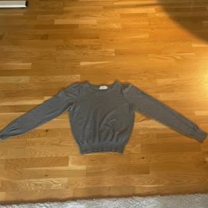 Grå stickad tröja från Minus - Snygg grå stickad tröja från Minus i storlek XS. Tröjan har rund halsringning, långa ärmar med lätt puff vid axlarna och ribbade muddar i nederkant och vid ärmslut. Perfekt för lager-på-lager och enkel att matcha med jeans eller kjol.