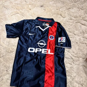 PSG Ronaldinho 21 Nike fotbollströja L - Klassisk PSG fotbollströja från Nike med Ronaldinho och nummer 21 på ryggen. Tröjan är marinblå med en röd vertikal rand, vita detaljer och PSG-logga på bröstet. Tillverkad i Dri-FIT material som andas. Kortärmad modell med krage och broderade loggor.