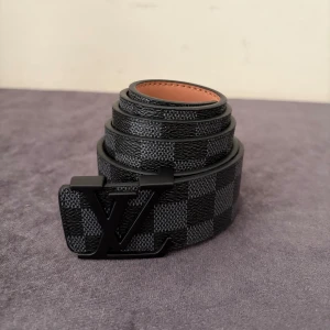 Louis Vuitton rutig svart bälte - Snyggt svart bälte från Louis Vuitton med klassiskt rutigt mönster och stor LV-logga i svart metall på spännet. Bältet är gjort i skinn med beige insida och har en exklusiv känsla. Perfekt för dig som vill ha en stilren accessoar med lyxig vibe.