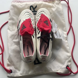Adidas Predator Elite FT S fotbollsskor - Adidas Predator Elite FT S fotbollsskor i vitt med svarta och röda detaljer. Skorna har snörning, täckt plös och mönstrad ovandel för extra grepp. Sulan har fasta dobbar för bästa grepp på planen. Perfekta för dig som vill sticka ut på matchen. Helt nya diskuterar gärna pris! 