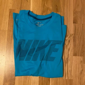 Blå Nike Dri-Fit t-shirt - Snygg blå t-shirt från Nike med stort svart logotryck över bröstet. Tillverkad i Dri-Fit-material som andas och håller dig torr. Klassisk passform med rund hals och korta ärmar, perfekt för träning eller chill.