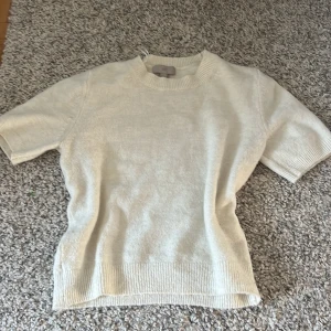 Beige stickad tröja från H&M - Säljer en beige stickad tröja från H&M i storlek S. Tröjan har korta ärmar, rund halsringning och ribbade muddar vid ärmslut och nederkant. Perfekt för lager-på-lager och enkel att matcha med olika outfits.
