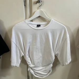 Vit t-shirt med snörning från Gina Tricot - Vit t-shirt i bomull från Gina Tricot med korta ärmar och cool snörningsdetalj i sidan. Avslappnad passform och rund halsringning, perfekt till jeans eller kjol.