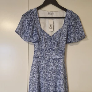 Blå blommig kortklänning med puffärm - Supersöt blå kortklänning med vitt blommönster och puffiga korta ärmar. Klänningen har v-ringning, knappar framtill och knytband i ryggen för extra snygg passform. Perfekt för sommarens alla tillfällen!