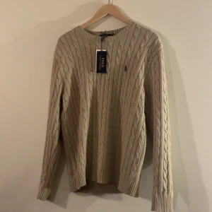  stickad tröja från Ralph Lauren - Säljer en stilren beige stickad tröja från Ralph Lauren helt ny med kabelstickat mönster. Tröjan har långa ärmar och en liten broderad logotyp på bröstet. Perfekt för en klassisk och tidlös look. Skriv vid frågor!!!