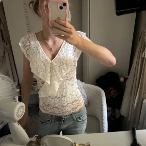spets body med volanger  - Supersöt vit body i spets med v-ringning och volangdetalj framtill. Ärmlös modell med lätt transparent känsla och blommigt spetsmönster. Perfekt att styla med jeans eller kjol. Strl S aldrig använd 