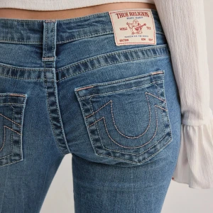 True Religion bootcut jeans blå - Säljer ett par blå bootcut jeans från True Religion med ikoniska bakfickor och ljusrosa kontrastsömmar. Jeansen har medelhög midja och klassisk femficksdesign. Materialet är mjukt denim i bomull med lätt stretch för skön passform. Nypris 1400!