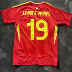 Spanien Lamine Yamal matchtröja Adidas - Röd Spanien fotbollströja från Adidas med gula detaljer, nummer 19 och Lamine Yamal på ryggen. Tröjan har korta ärmar, gula ränder på axlarna och Spaniens landslagsemblem på bröstet. Tillverkad i lätt och ventilerande material, perfekt för fotbollsfans.