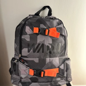 Grå camo ryggsäck från Warp - En ryggsäck i fint skick. Perfekt till skolstart eller om man behöver något extra att packa i. Praktiska spännen på utsidan och ett dator fack inuti.