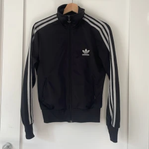Svart vintage Adidas track jacket med ränder - Svart vintage Adidas track jacket med klassiska vita ränder längs ärmarna och stort logotryck på ryggen. Jackan har dragkedja framtill, ståkrage och ribbade muddar. Perfekt för en sportig och avslappnad stil.