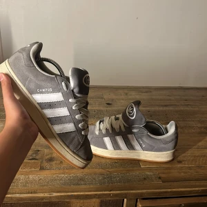 Grå Adidas Campus sneakers i mocka - Snygga grå Adidas Campus sneakers med klassiska vita ränder och logga på plösen. Ovandel i mjuk mocka, vita snören och beige sula med gummidetaljer. Perfekta för dig som gillar retrostil och streetwear.