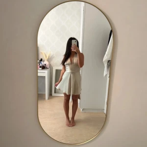 Champagnefärgad omlottklänning i satin - Supersnygg kort klänning i champagnefärgad satin med omlott och djup v-ringning. Klänningen har breda axelband som knyts i nacken och markerad midja. Perfekt för dig som vill ha en elegant och trendig look med lite extra shine.