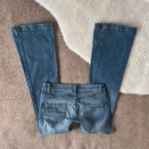 Blå bootcut jeans från next  - Snygga blå jeans med klassisk bootcut-modell och fem fickor. Jeansen har en låg midja och raka ben som blir vidare nedtill. Tillverkade i mjukt denimtyg med diskreta slitningar och detaljerade bakfickor. Midjemått- 38 innerbenslängd- 75