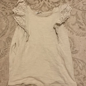 Vit topp med volangärmar från H&M - Supersöt vit topp från H&M med broderade volangärmar i spets. Ärmlös modell med rund halsringning och detaljerade hålmönster på axlarna. Perfekt för dig som gillar romantiska och luftiga plagg med extra detaljer.