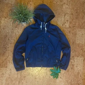 Blå vindjacka från Polo Ralph Lauren - Snygg marinblå vindjacka från Polo Ralph Lauren med huva och vita snören. Jackan har dragkedja framtill, två fickor med dragkedja och elastiska muddar. Liten röd logga broderad på bröstet. Perfekt för blåsiga dagar och enkel att matcha.