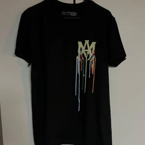 Svart Amiri t-shirt med tryck - Svart t-shirt från Amiri med coolt tryck framtill av droppande färg och stort grafiskt tryck på ryggen med texten AMIRI och en hand som håller en klot. Klassisk passform och korta ärmar, perfekt för dig som gillar streetwear med edge.