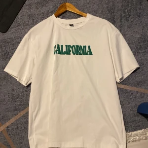 Vit t-shirt med grön text från SHEIN - Vit t-shirt från SHEIN i storlek M med coolt grönt tryck 'CALIFORNIA' på bröstet. Klassisk passform med rund hals och korta ärmar. Perfekt för en avslappnad streetstyle-look.