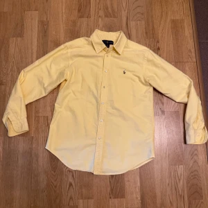 Gul skjorta från Ralph Lauren - Snygg gul skjorta från Ralph Lauren med klassisk krage och broderad logga på bröstet. Skjortan har lång ärm, knäppning framtill och är gjord i mjuk bomull. Perfekt för dig som vill ha en stilren och fräsch look.