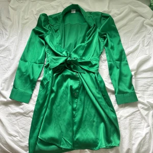 Grön omlottklänning från Lindex - Snygg grön kortklänning från Lindex i glansigt material. Klänningen har långa ärmar, omlottmodell med knyt i midjan och klassisk krage. Perfekt för dig som gillar färg och vill sticka ut lite extra.