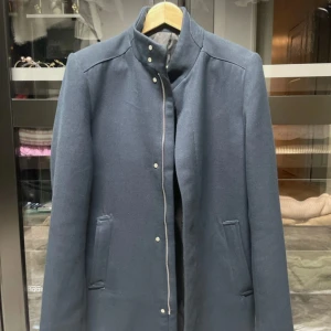 Mörkgrå rock med ståkrage - Snygg mörkgrå rock med minimalistisk design och ståkrage. Jackan har raka linjer, dolda knappar och två fickor framtill. Perfekt för dig som gillar stilrena och klassiska plagg.