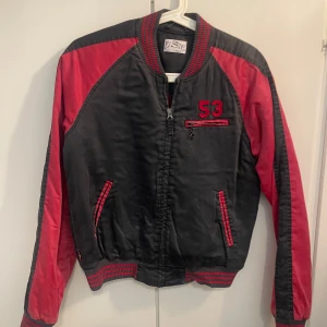 Levi's svart och röd bomberjacka S - Snygg svart och röd bomberjacka från Levi's med broderad text på ryggen och siffran 53 framtill. Jackan har dragkedja, randiga muddar och röda detaljer på fickor och axlar. Perfekt för dig som vill sticka ut med en sportig och retro vibe.