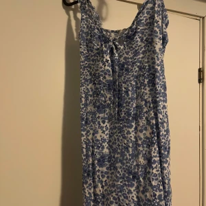 Blåvit blommig klänning från Nelly - Supersöt blåvit klänning från Nelly med blommigt mönster, knytning framtill och dragkedja i ryggen. Lätt och luftig modell som passar perfekt till varma dagar. Ärmlös design och smickrande passform.