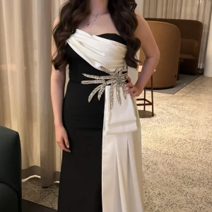 Svart och vit långklänning med drapering - Säljer en elegant svart och vit långklänning med axelbandslös design och draperad vit detalj över bysten. Klänningen har en glittrig dekoration i midjan och ett långt vitt tygstycke som faller ner längs sidan. Perfekt för dig som vill sticka ut på bal eller fest. Den är använd en gång! 