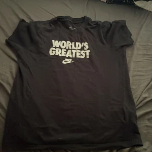 Svart Nike t-shirt med tryck - Svart t-shirt från Nike med stort vitt tryck på bröstet där det står 'WORLD'S GREATEST' och den klassiska Nike-loggan under. Klassisk passform med rund hals och korta ärmar. Perfekt för dig som vill ha en clean och sportig stil.