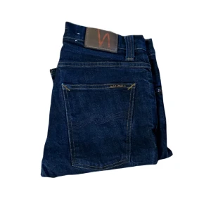 Nudie Grim Tim jeans - Snygga mörkblå jeans från Nudie Jeans med klassisk femficksdesign och kontrastsömmar. Jeansen har raka ben och är tillverkade i slitstarkt denim med Nudie-logga på bakfickan. Perfekta för dig som gillar stilrena och tidlösa jeans. Är lite osäker på om de kan vara uppsydda då jag tycker de är någon cm kort för att vara L32. KAN EJ HA DENNA STORLEK. Midja 39cm så de är väldigt små i midjan för sin storlek och så passar mer som W30 men på etiketten står det W34!