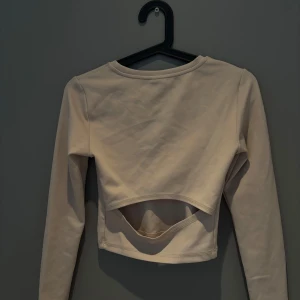 Beige croppad topp från Gina Tricot - Säljer en beige långärmad croppad topp från Gina Tricot. Toppen har en cool cut out-detalj framtill som ger en trendig look. Träningströja. 