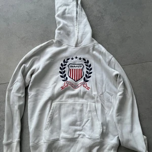 Vit hoodie från GANT med broderad logga - Vit hoodie från GANT med stor broderad logga och emblem i marinblått och rött på bröstet. Klassisk känguruficka framtill, mjuk insida och huva.  