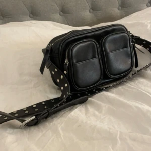 Svart axelväska med nitar och dragkedja - Cool svart axelväska i skinnimitation med bred axelrem dekorerad med silvriga nitar. Väskan har två framfickor med dragkedja och ett större huvudfack. Perfekt för dig som vill ha en praktisk väska till vardags. Den har en sminkfläck inuti men som ny utanpå! Pris kan diskuteras