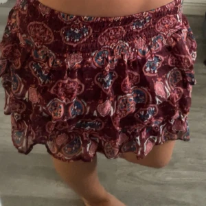 Vinröd volangkjol med mönster - Supersöt kort kjol i vinrött med färgglatt paisleyinspirerat mönster i rosa, blått och orange. Kjolen har bred resår i midjan och flera volanger som ger en flowy och lekfull känsla. Perfekt för varma dagar när du vill ha något bekvämt och snyggt. Är storlek 152 men passar mig som bär storlek s!!