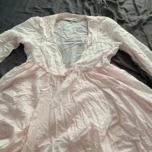 Ljusrosa långärmad klänning från Love & Pie - Superfin ljusrosa klänning med lång ärm, djup v-ringning och broderade detaljer. Klänningen har markerad midja och är gjord i ett tunt, luftigt bomullstyg. Perfekt för sommarens varma dagar eller när du vill känna dig extra söt.