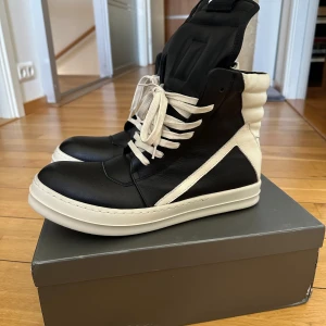 Svartvita high-top sneakers med dragkedja - Säljer ett par Rick owen geobasket high-to sneakers med cool design och kraftig plös. Skorna har både snörning och dragkedja på sidan för enkel påtagning. Tjock vit sula och detaljerad paneldesign. Perfekta för dig som vill sticka ut med din stil. Passar 45/46