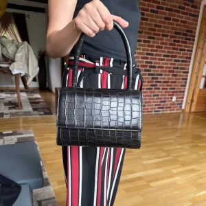 Svart croco handväska med axelrem - Snygg svart handväska i croco-mönstrat skinnimitation. Väskan har både ett stadigt handtag och en justerbar axelrem för olika stilar. Stilren och boxig form med lock och guldfärgade detaljer. 