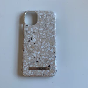 Beige terrazzo mobilskal iDeal of Sweden  - Snyggt mobilskal från iDeal of Sweden med terrazzo-inspirerat mönster i beige, vitt och inslag av svart. Skalet har en glansig finish och är gjort i slitstark syntet. Perfekt för dig som vill ha en trendig och stilren look till mobilen. Men är lite trasig i högra hörnet längst ner men funkar bra 🤗